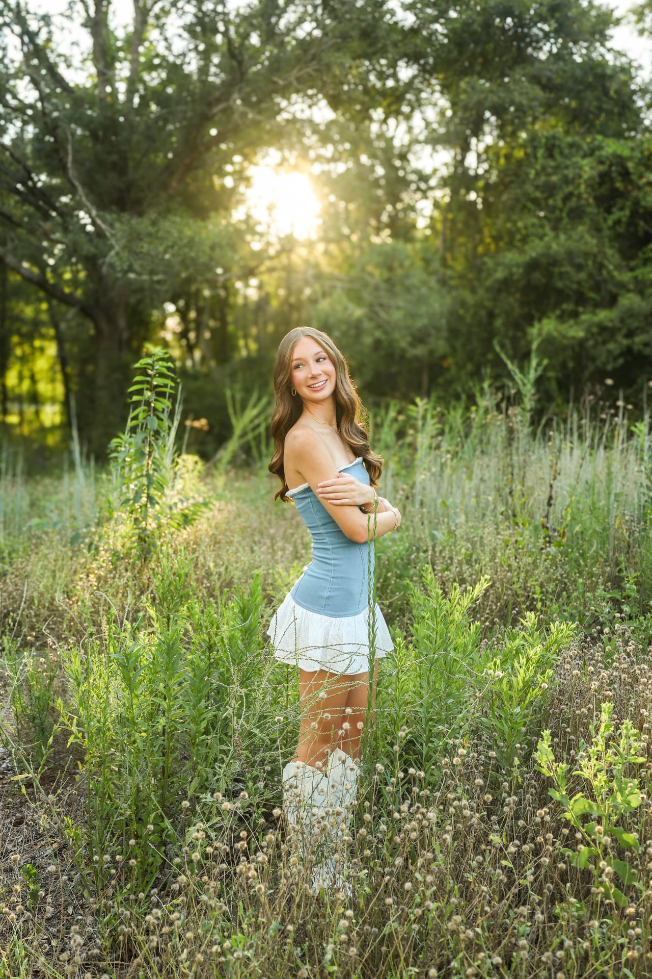 Senior in light blue dress and cowboy boots in tall grass at sunset