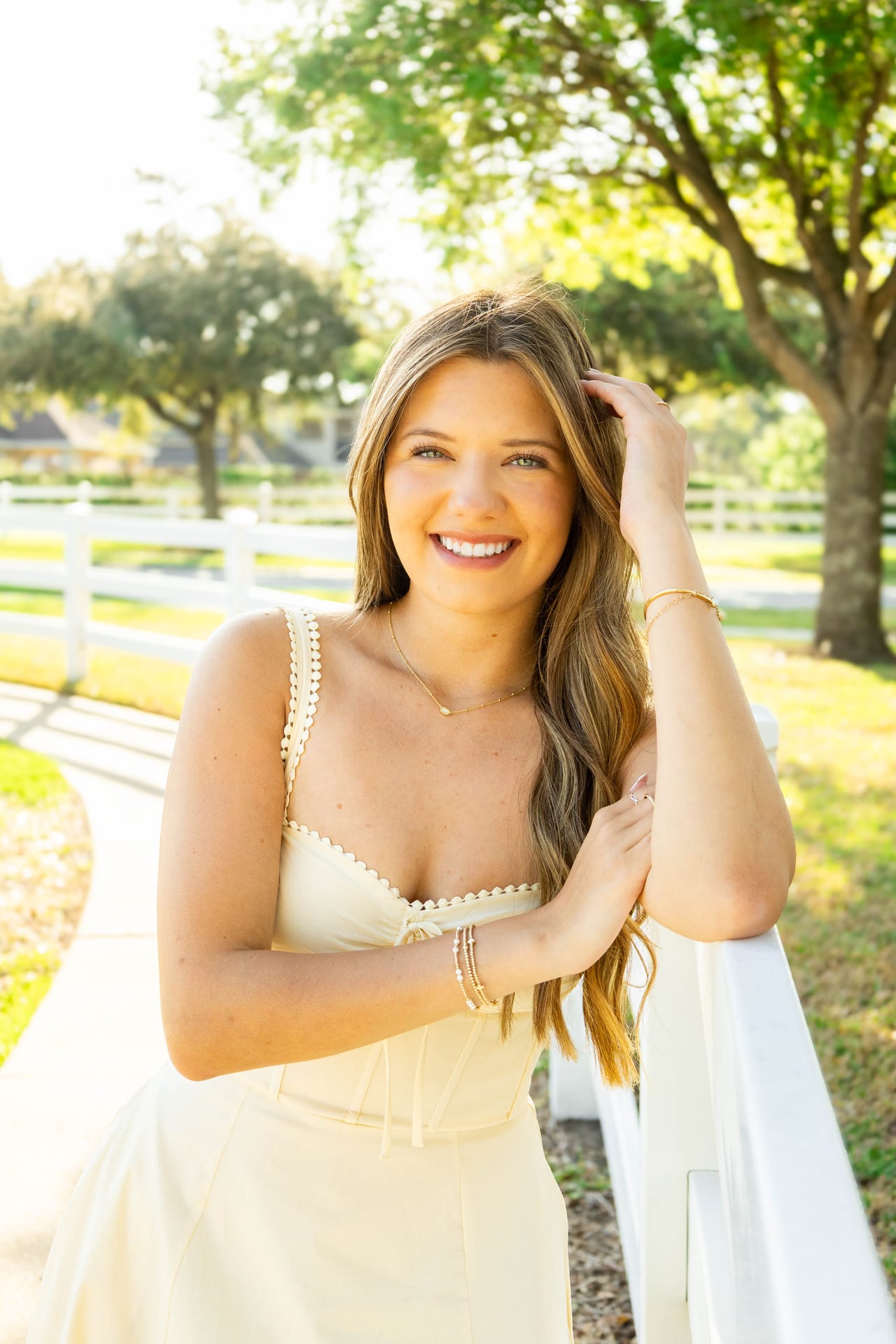 Senior in cream dress leaning on a white fence at golden hour