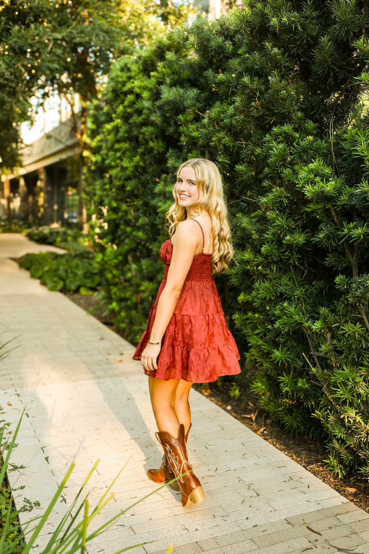 Senior in red dress and cowboy boots walking past green hedges