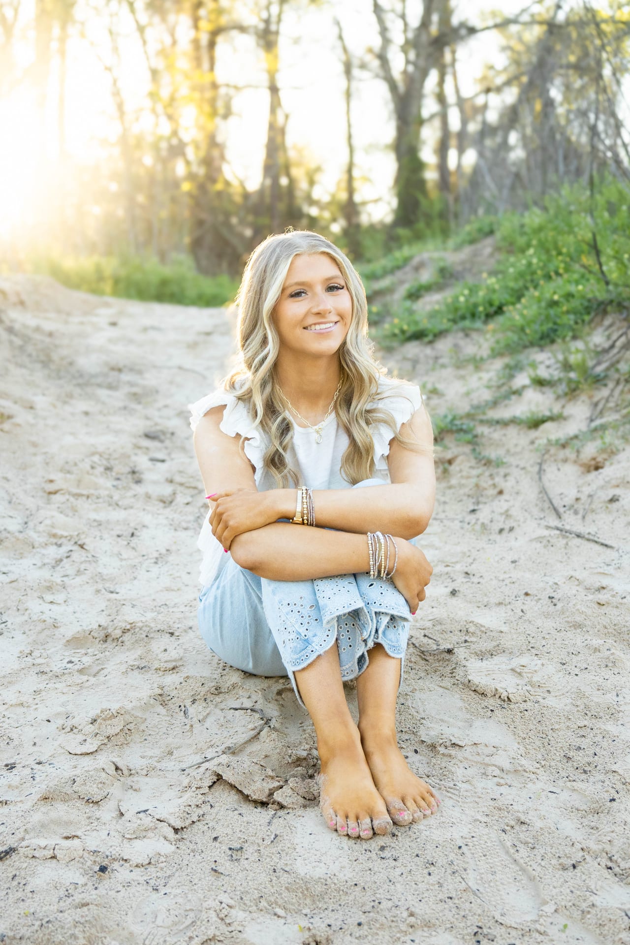 Senior in light blue dress sitting barefoot on sand at golden hour